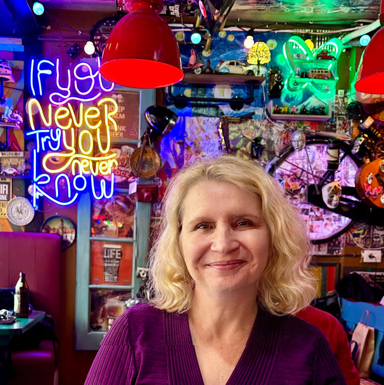 In Tirana Albania. Photo by author. Photo of a woman with short wavy blond hair facing the camera and smiling in a restaurant full of bright neon lights including on with the sign If You Never Try You Never Know.